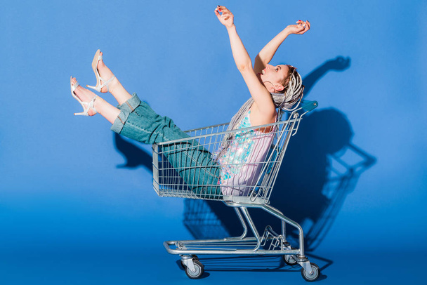 mooi stijlvol meisje in winkelwagen gebaren op blauw  - Foto, afbeelding