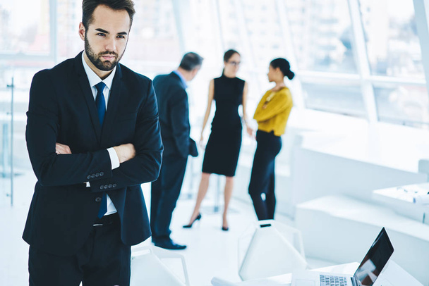 Confident man if elegant suit looking away while male and female colleagues on blurred background communicate about trade and accounting, serious bearded businessman thinking about idea for project - Photo, Image
