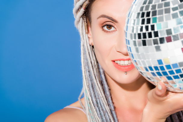 beautiful girl with dreadlocks covering face with disco ball isolated on blue  - 写真・画像