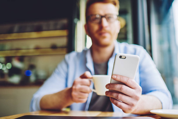 Selective focus on male hand holding cellular phone and looking away while waiting for updating of application during coffee time indoors, man using 4g wireless internet on modern mobile gadget - Fotoğraf, Görsel