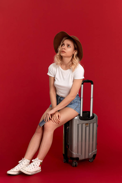 Bir günbatımı şapka blod kıvırcık saçlı endişeli genç kadın kırmızı bir arka plan önünde pasaport tutan gri bir bagaj çantası üzerinde oturur - Fotoğraf, Görsel