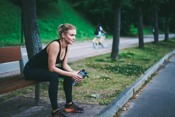 Caucasian female runner taking rest on wooden bench with aluminium thermos in hands, active sporty girl in black tracksuit looking away and feeling good from well done hard training on nature - Foto, Imagen