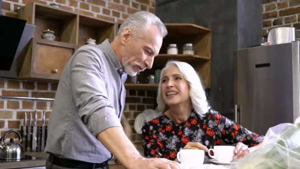 Cheerful lovely elderly couple sitting together while talking and drinking coffee at home - Imágenes, Vídeo