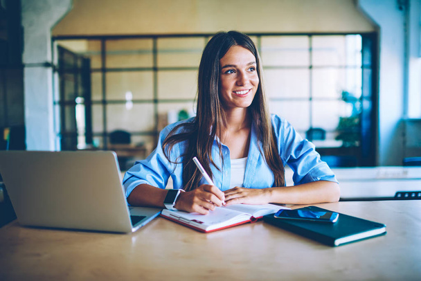 Smiling successful woman with netbook enjoying planning process while sitting at desktop with modern laptop device indoors, young positive female student thinking about ideas for school project - Photo, image