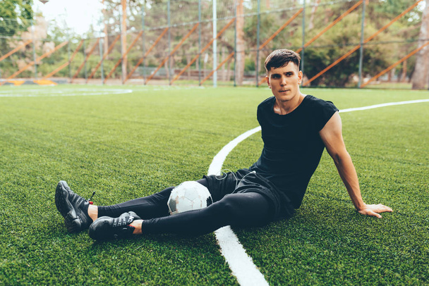 Handsome young soccer player man taking a rest after training session outdoors. Healthy and fit man sitting and relaxing at green lawn after workout exercises. People and sport concept - Photo, image