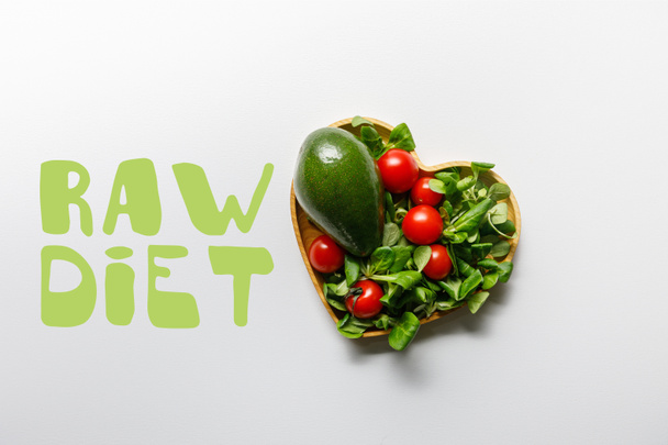 top view of fresh green vegetables in heart shaped bowl on white background with raw diet lettering - Fotó, kép