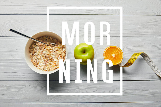 flat lay with breakfast cereal in bowl, apple, orange and measuring tape on wooden white background with morning lettering - Foto, immagini