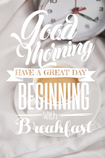 Top uitzicht van koffie in witte kop op schotel in de buurt van zilveren wekker op beddengoed met Goedemorgen, hebben een geweldige dag beginnen met ontbijt belettering - Foto, afbeelding