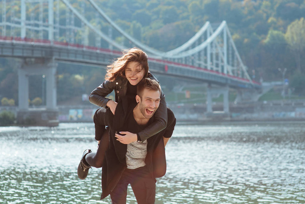 beautiful amazing funny cheerful young couple (man and woman) outdoor by the river on bridge background. Girlfriend riding on boyfriend back. Family, love and friendship concept - Photo, Image