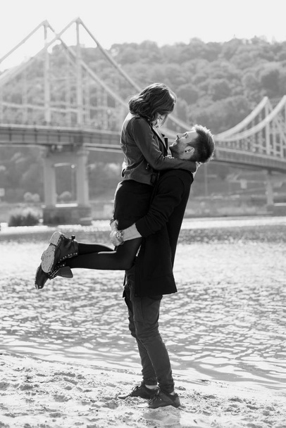 nádherný úžasný veselý mladý pár (muž a žena) venku u řeky na pozadí mostu. Přítelkyně jezdila s přítelem. Koncepce rodiny, lásky a přátelství - Fotografie, Obrázek