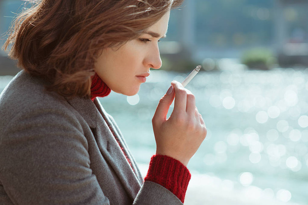 hermosa mujer joven con estilo triste estresante en suéter y abrigo fumar cigarrillo cerca del río
 - Foto, Imagen