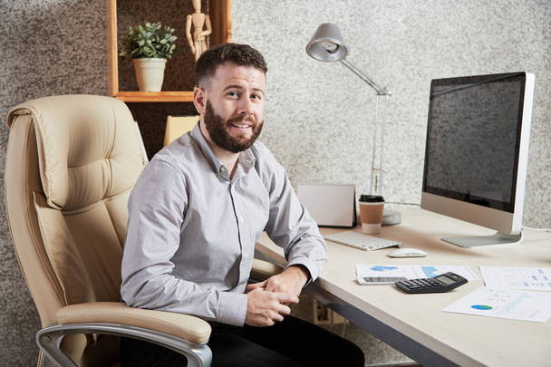 Portréja sikeres szakállas férfi vezető ül kényelmes szék közelében a munkahelyen, és mosolyogva a kamera - Fotó, kép