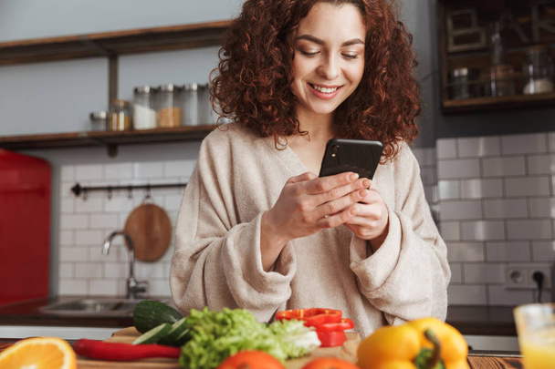 Photo of happy caucasian woman holding smartphone while cooking  - Photo, Image
