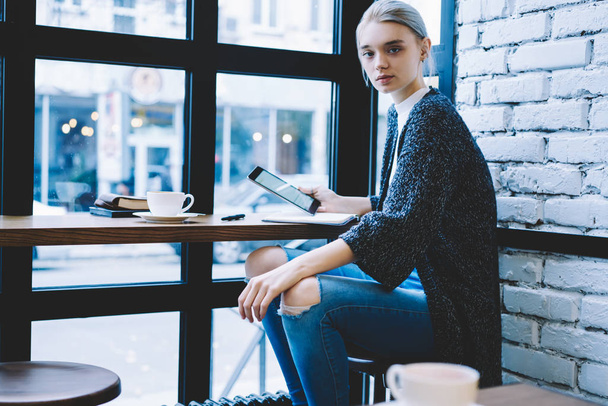 Pretty modern blonde hair female sitting at table near window in coffee shop and holding digital tablet. Millennial hipster girl spending recreation time using technology  - Photo, image