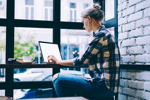 Side view of young blonde hair female in casual clothes using mock up laptop while studying at table in modern coffee shop interior sitting near window. Young contemporary woman working on freelance - Zdjęcie, obraz