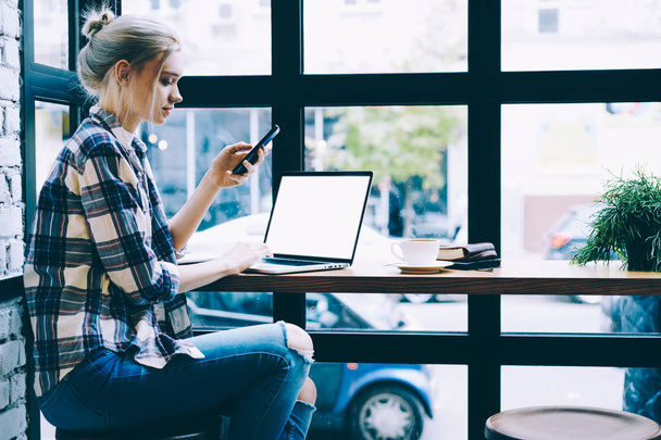 Modern sarışın saç kadın masada oturan cep telefonu uygulama üzerinden Laptop bilgisayar tarama ağı Mock kadar yakın. Millennial hipster kız harcama zaman kullanarak teknoloji içinde kahve dükkanı - Fotoğraf, Görsel