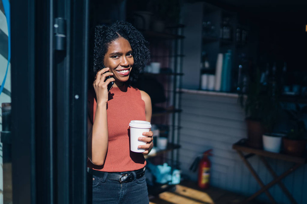Half length portrait of cheerful millennial hipster girl with hot drink in hand looking at camera with candid smile on face and enjoying communication via smartphone application with best friend - Φωτογραφία, εικόνα