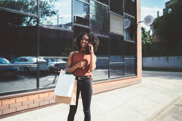 Cheerful hipster girl holding packages bags with purchases after shopping and calling for boast of catch good sales in mall,positive dark skinned female shopaholic with paper bags enjoying mobile talk - Fotó, kép