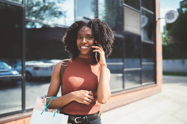 Portrait of carefree dark skinned female shopaholic with shopping bags in hand calling to friend for communicating about sales connected to roaming internet on cellphone, concept of Black Friday - 写真・画像