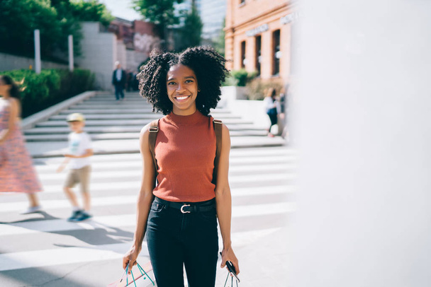 Half length portrait of cheerful female teenager dressed in casual wear standing on urban setting and smiling at camera during sunny summer weather in city, concept of young dark skinned millennial - Fotó, kép