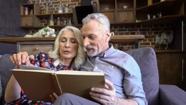 Pleased lovely elderly couple reading together while sitting together on sofa at home - Imágenes, Vídeo