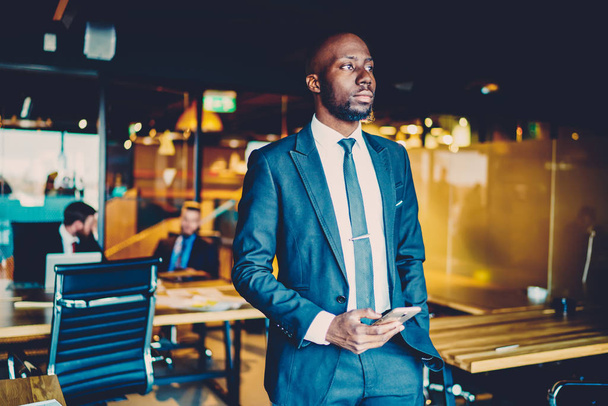 Overpeinend African American zakenman gekleed in formele slijtage staande met smartphone in kantoor interieur. Doordachte Dark gevild trotse CEO met mobiele telefoon in de handen van de planning van zijn werkdag - Foto, afbeelding