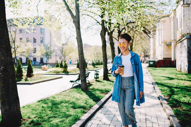 Half length portrait of cheerful female teenage tourist with takeaway cup walking around spring city, positive african hipster girl in stylish jeans jacket looking at camera enjoying sunny weather - Photo, Image