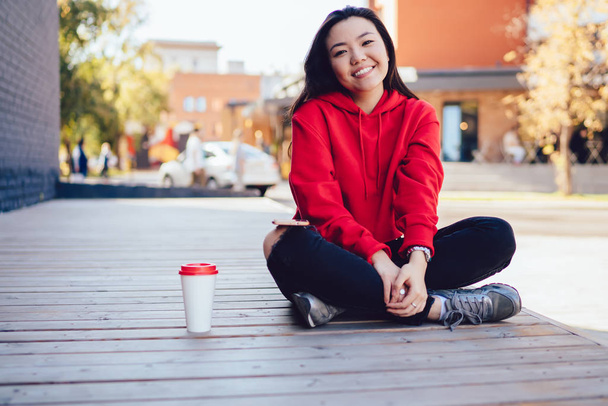 Portrait of cheerful female teenager dressed in stylish casual look taking rest on urban setting during sunny day, positive asian hipster girl sitting near takeaway cup with copy space area - Фото, изображение