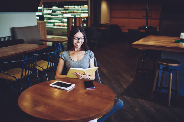 Happy female teenager reading romantic novel in coffee shop feeling prosperous from good story, positive caucasian hipster girl in spectacles smiling while spending time for literature hobby - Foto, immagini