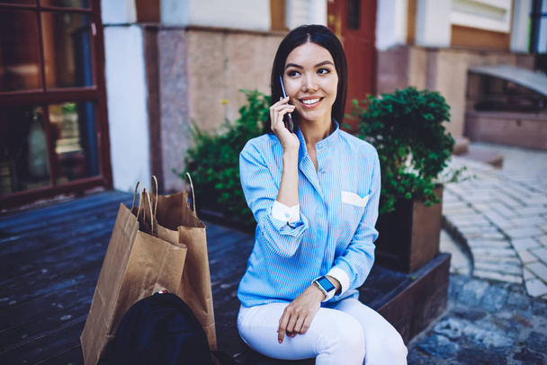 Successful female customer enjoying resting on urban setting after spending time in male for shopping and buying new trendy clothes, happy woman making telephone conversation via app outdoors - Фото, изображение