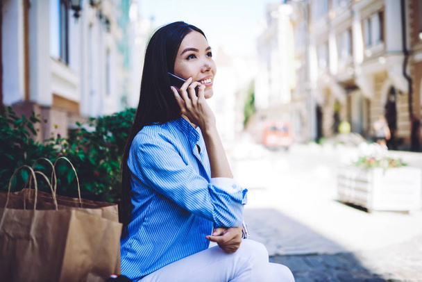 Young positive female shopaholic smiling and calling to taxi customer service for rent car using roaming 4g internet for communicate outdoors, happy hipster girl talking with friend via smartphone - Photo, image