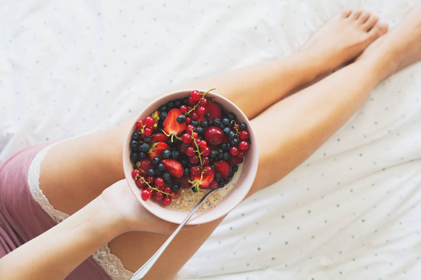 Terveellinen kesäaamiainen sängyssä. Nainen syö kaurapuuroa marjoilla. Terveellistä syömistä. Yläkuva
 - Valokuva, kuva