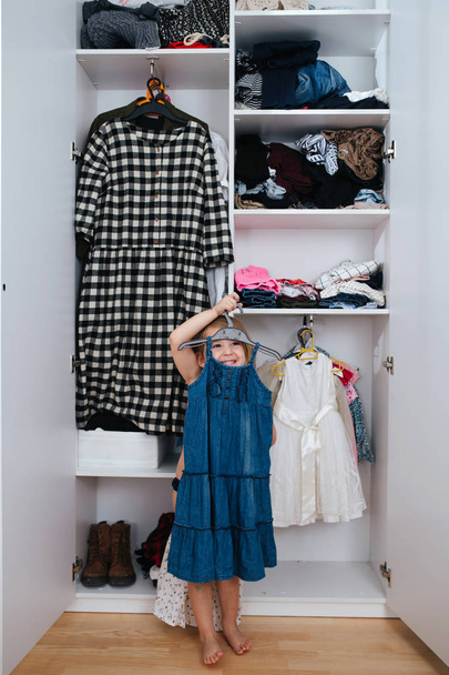 Little jolly girl trying on new dress in front of open wardrobe full of clothes - Фото, зображення