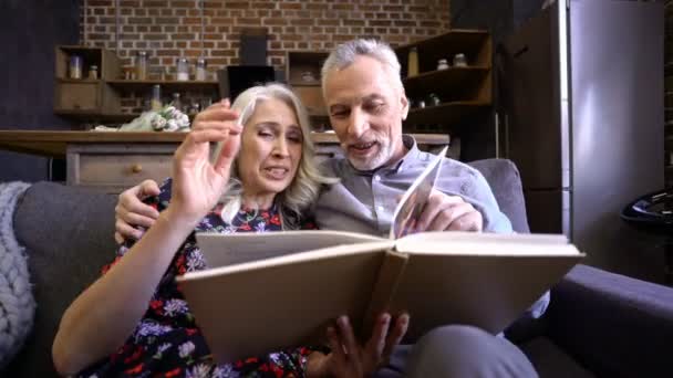 Cheerful elderly elegant couple sitting together on sofa and reading book at home while man kissing woman - Video