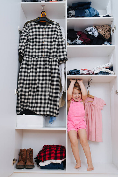 Portrait of little girl in new pink overalls playing inside of open wardrobe - Foto, afbeelding