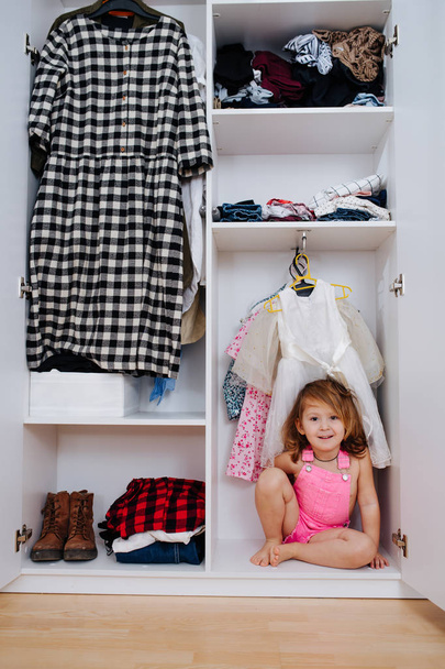 Portrait of little girl in new pink overalls sitting inside of open wardrobe - Fotoğraf, Görsel