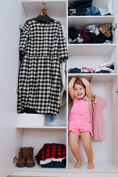 Portrait of little girl in new pink overalls playing inside of open wardrobe - Photo, image