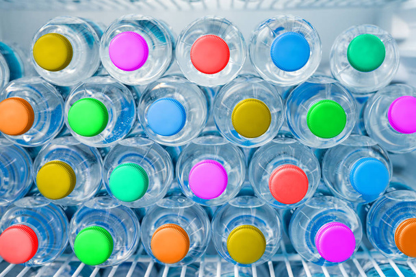 Σειρές από πολλές διαφανείς πλαστικές φιάλες πόσιμου νερού με φωτεινά πολυλόφυλλα πολύχρωμα καπάκια σε λευκό ψυγείο. Αποθηκευτική στοίβα μεταλλικού νερού στο ψυγείο. Πρόληψη της υγειονομικής περίθαλψης και της αφυδάτωση - Φωτογραφία, εικόνα