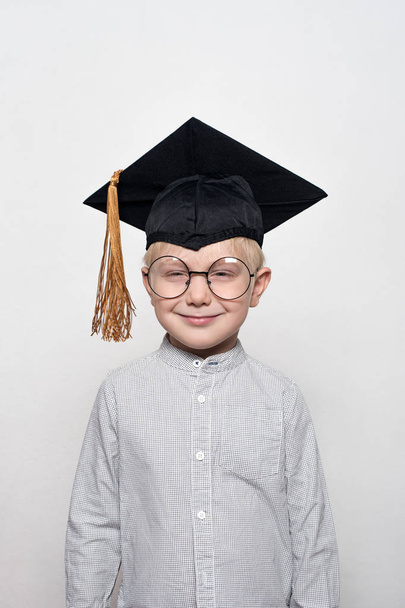 Portret cute blond chłopca w dużych okularach i kapelusz akademicki. Białe tło. - Zdjęcie, obraz