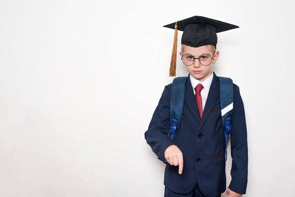 Seriózní školák v obleku, brýle a akademický klobouk míří dolů. Školní koncept. Bílé pozadí. - Fotografie, Obrázek