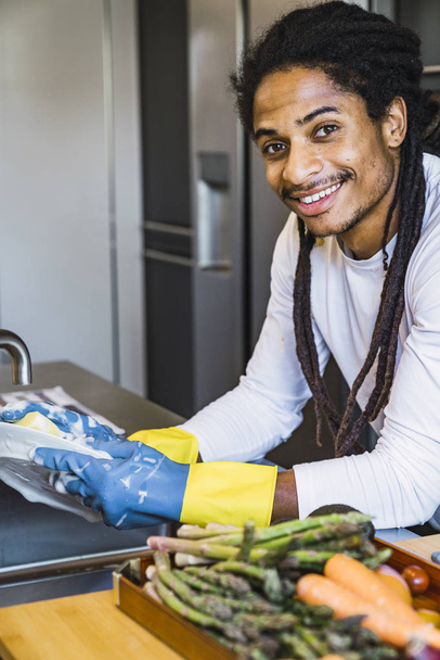 Καθαρισμός χεριών. Νέος μαύρος άντρας που πλένει πιάτα στη μοντέρνα κουζίνα. Μια νεαρή γυναίκα που πλένει πιάτα χειροκίνητα με το χέρι, φορώντας μπλε και κίτρινα ελαστικά γάντια καθαρισμού. Κουράστηκα να καθαρίζω. - Φωτογραφία, εικόνα