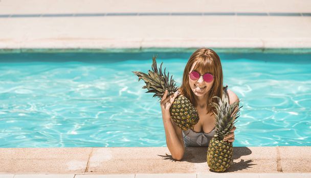Caucasian redheaded girl with the body inside the water in the pool grabbed at the edge of the pool with two pine cones in her hands. Sticking out her tongue with sunglasses with red lenses - Foto, Imagen