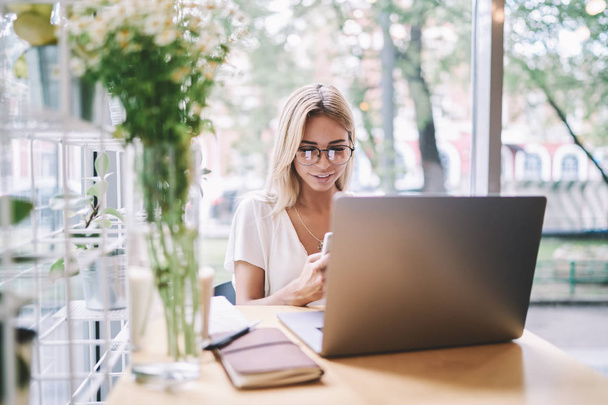 Smiling woman in eyewear updating notification on smartphone application for chatting while sitting at cafeteria with laptop device, millennial hipster girl enjoying good wifi connection on cellular - Фото, изображение