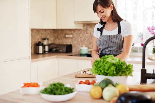 Beautiful smiling dedicated Caucasian brunette in apron standing in kitchen and chopping mushrooms. On table are lots of vegetables. Cooking at home concept. - Foto, afbeelding