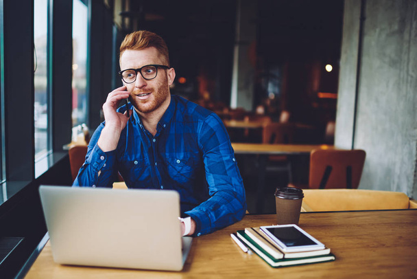 Successful male freelancer calling to customer service for discussing antivirus application and notification for on laptop computer, hipster guy making positive smartphone conversation indoors - Photo, Image