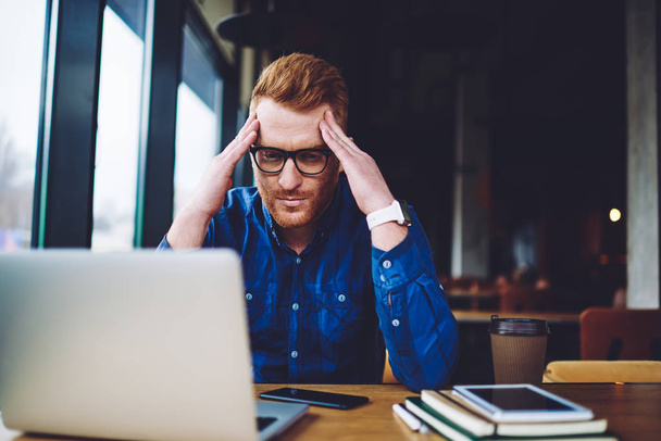 Puzzled man in optical spectacles pondering on problem with program code for creating new application for communication via laptop computer, young hipster guy feeling headache during distance job - Photo, Image