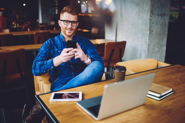Portrait of positive male blogger in eyewear sitting with cellular phone in hand and smiling in camera during break from working remotely on modern netbook, concept of generation and communication - Zdjęcie, obraz