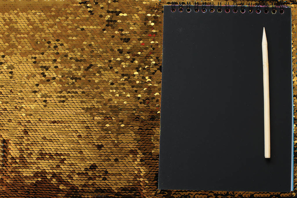 空の黒いシートと木製のスクレーパーを持つノートブックは、金のスパンコールの明るい光沢のある背景にあり、手紙を書くスペース、黒い紙にテキストを書きます - 写真・画像