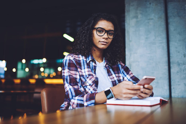 Portrait of attractive african female student sitting at table with cellphone and looking at camera during break from studying in university campus, dark skinned hipster girl using technology - Фото, зображення