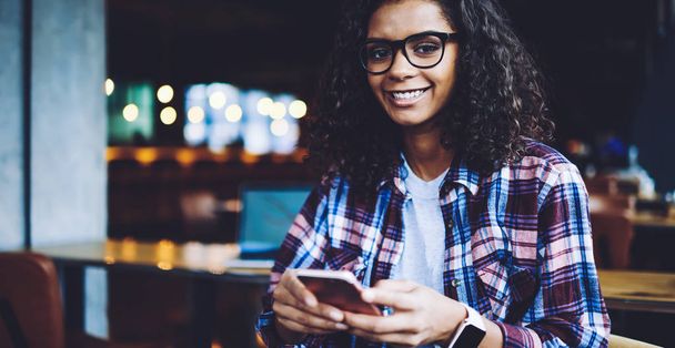 Portrait of cheerful black woman in eyewear holding cellular gadget in hand and smiling during leisure time, happy millennial female blogger feeling good from received email form follower via mobile - 写真・画像